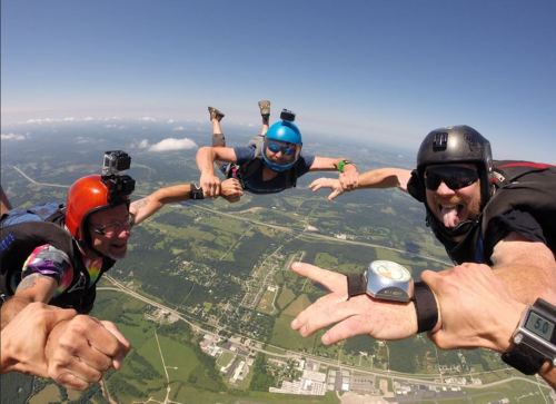 Three skydivers in freefall, smiling and posing for the camera against a clear blue sky and green landscape below.