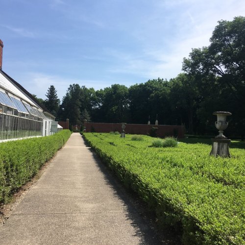 A pathway leads through neatly trimmed hedges to a greenhouse, surrounded by lush greenery and trees.