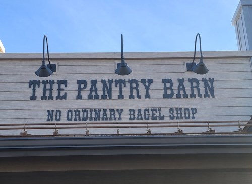 Sign for "The Pantry Barn" with the tagline "No Ordinary Bagel Shop" against a clear blue sky.