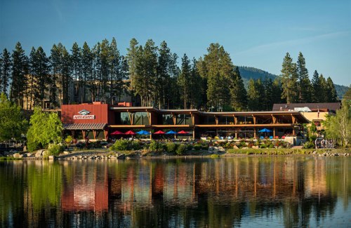 A lakeside lodge surrounded by trees, featuring outdoor seating and colorful umbrellas, reflecting in the water.