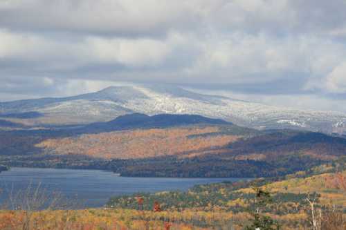 A scenic view of a lake surrounded by autumn foliage and mountains, with some snow on the peaks under a cloudy sky.