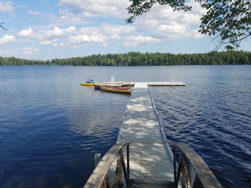 A wooden dock extends into a calm lake, with colorful boats moored nearby and trees lining the shore under a blue sky.