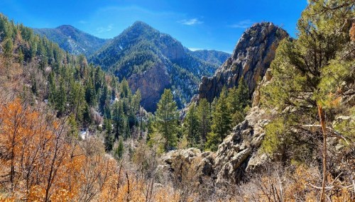 A scenic mountain landscape with lush green trees, rocky formations, and a clear blue sky.
