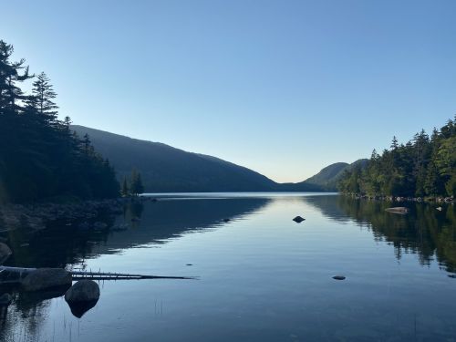 A serene lake surrounded by mountains and trees, reflecting the clear blue sky at dawn.
