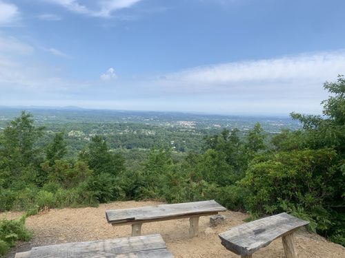 Scenic view from a mountain overlook, featuring benches and lush greenery under a blue sky with scattered clouds.