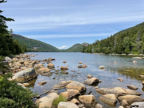 A serene lake surrounded by mountains and trees, with large rocks scattered along the shore under a blue sky.