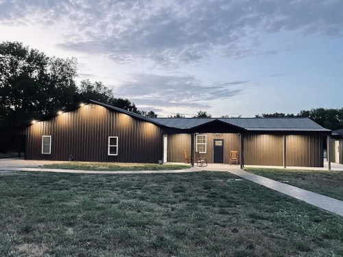 A modern, dark metal-clad house at dusk, with a grassy yard and a pathway leading to the entrance.