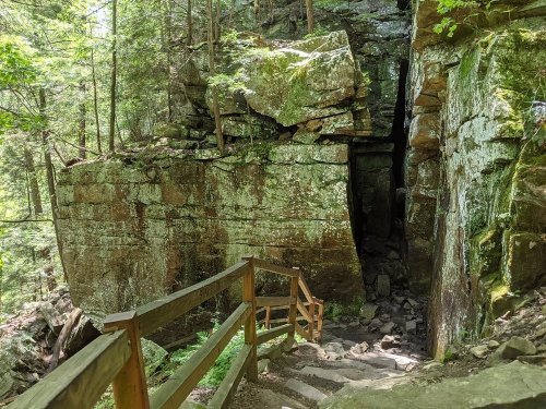 A wooden staircase leads down through a rocky, moss-covered forest with tall trees and a narrow crevice in the rocks.