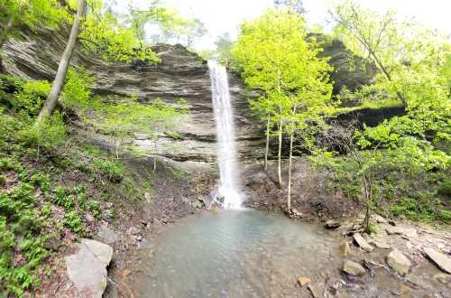 A serene waterfall cascades down rocky cliffs, surrounded by lush green trees and a calm pool below.