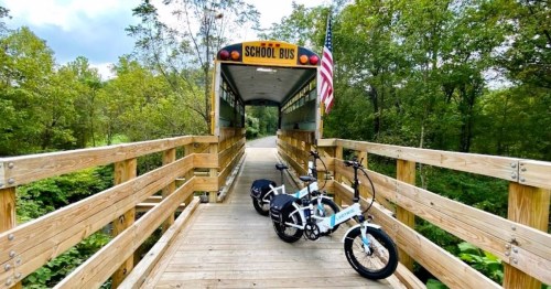 A wooden bridge with a school bus at one end and two bicycles parked beside it, surrounded by greenery.