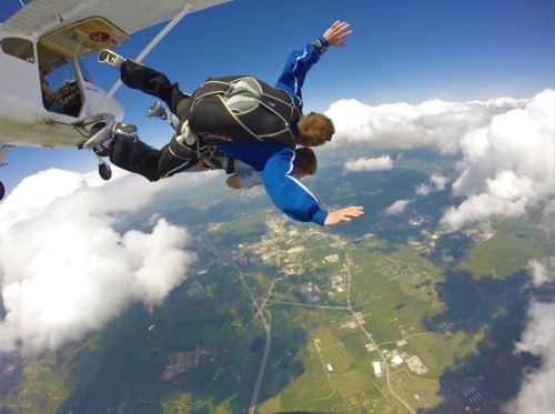 Two skydivers leap from a plane, arms outstretched, above a landscape of clouds and greenery.