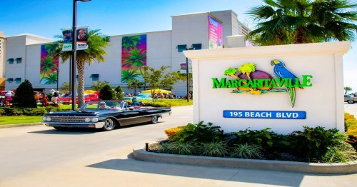 A classic car drives past the Margaritaville sign on Beach Blvd, with colorful buildings and palm trees in the background.