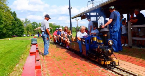 A miniature steam train with passengers departs from a station, surrounded by green grass and trees on a sunny day.