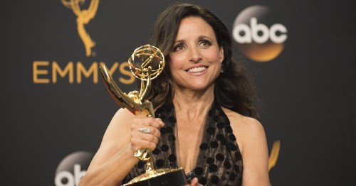 A woman in a black dress holds an Emmy award, smiling at a press event with a backdrop featuring the Emmy logo.