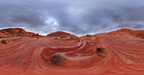 A panoramic view of red rock formations with swirling patterns under a cloudy sky.