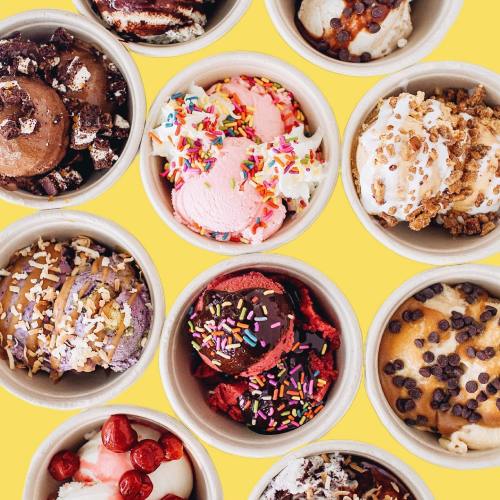 A colorful assortment of ice cream scoops in cups, topped with sprinkles, chocolate, and various toppings on a yellow background.