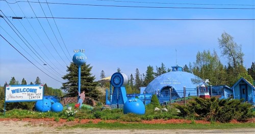 A quirky blue building with a dome, surrounded by blueberry-themed decor and signs for a heritage center and pies.