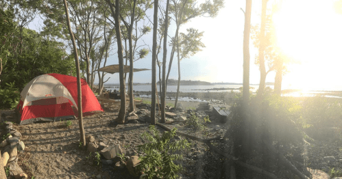 A red tent set up by a serene lakeside, surrounded by trees and a sandy area, with sunlight filtering through.
