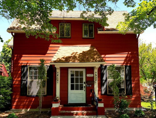 A charming red two-story house with a white door, black shutters, and surrounded by greenery on a sunny day.