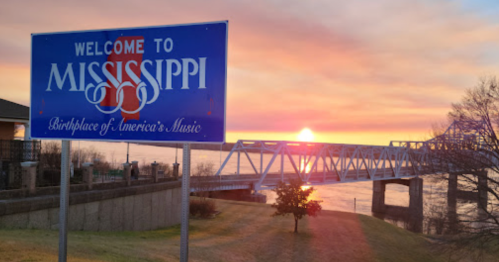 A welcome sign for Mississippi at sunset, with a bridge and river in the background.