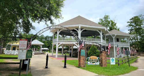 A charming farmers market pavilion with a large roof, surrounded by greenery and American flags, in a park setting.