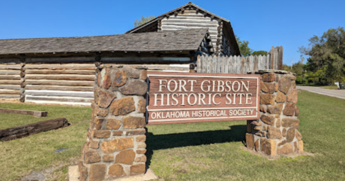 Sign for Fort Gibson Historic Site, Oklahoma Historical Society, in front of a log cabin structure on a clear day.