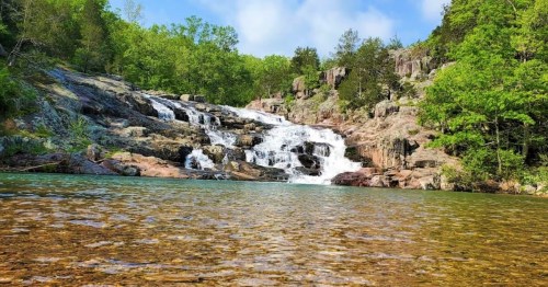 A serene waterfall cascades over rocks into a clear pool, surrounded by lush green trees and a blue sky.