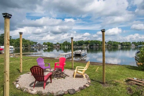 A serene lakeside scene featuring colorful chairs around a fire pit, with docks and trees in the background under a cloudy sky.