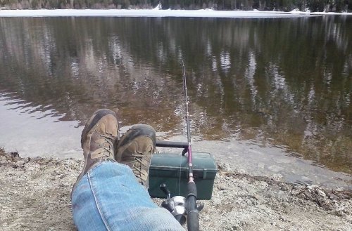 A person relaxes by a calm lake, feet resting on the shore, with a fishing rod and tackle box nearby.