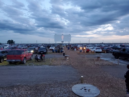 A drive-in movie scene with cars parked in a field, a large screen displaying a film, and people sitting on lawn chairs.