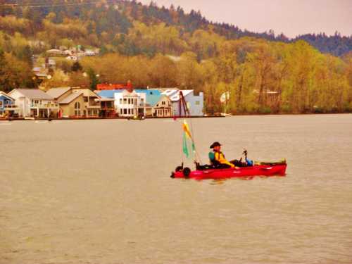 A person in a kayak paddles on a calm river, with colorful houses lining the shore in the background.