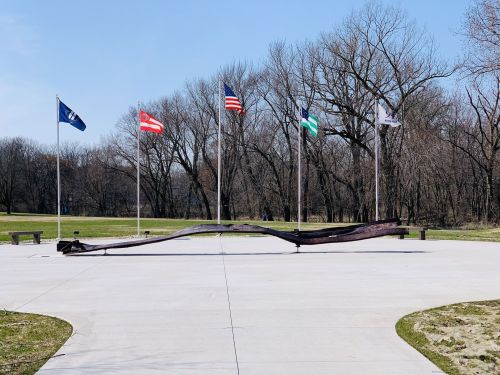 A sculpture on a concrete plaza with several flags flying in the background and bare trees lining the area.