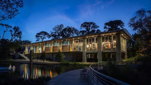 Modern building with large windows, surrounded by trees, reflecting in a calm water body at dusk.