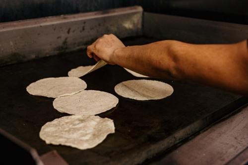 A hand flips tortillas on a hot griddle, with several tortillas cooking on the surface.
