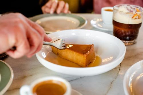 A hand using a fork to cut into a slice of flan on a white plate, with coffee cups and a glass of beer in the background.