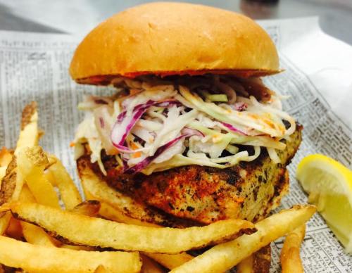 A grilled chicken sandwich topped with coleslaw, served with crispy fries and a lemon wedge on the side.