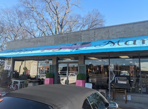 A storefront with a blue awning featuring the name "Manor," showcasing various items inside and a convertible parked outside.