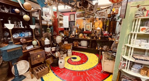 A cozy antique shop filled with various vintage items, furniture, and a colorful spiral-patterned rug on the floor.