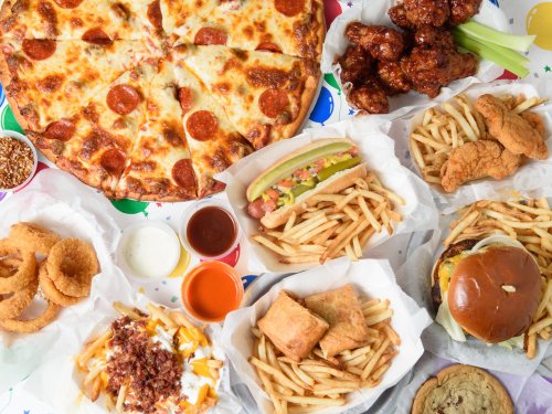 A spread of various fast foods including pizza, wings, fries, a hot dog, a burger, and onion rings on a colorful table.