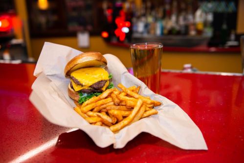 A cheeseburger with lettuce and fries on a red countertop, alongside a glass of beer in a bar setting.