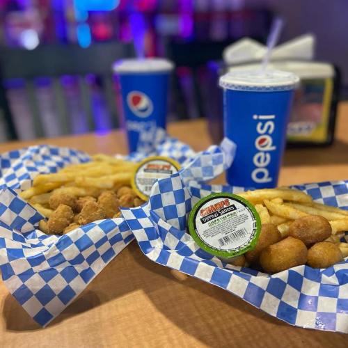 Two plates of food with chicken bites, fries, and hushpuppies, accompanied by Pepsi drinks on a checkered table.