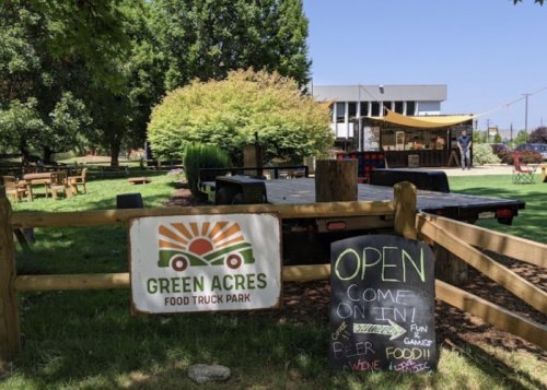 A sunny food truck park with a sign reading "Green Acres" and a chalkboard announcing it's open for food and fun.