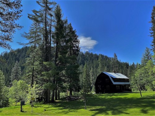 A black house surrounded by tall trees and lush green grass, set against a backdrop of mountains and a clear blue sky.