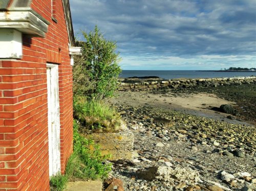 A red brick building near a rocky shoreline under a cloudy sky, with water visible in the background.