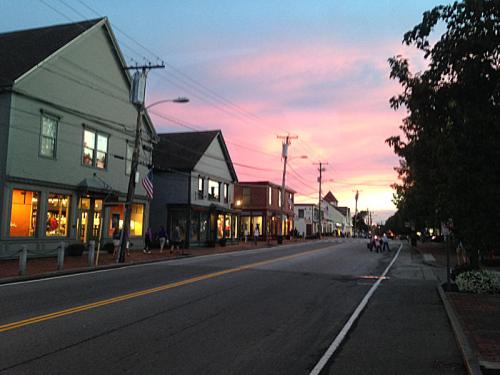 A quiet street lined with shops under a colorful sunset sky, with trees and power lines in the background.