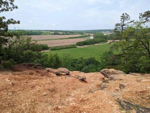 A scenic view from a rocky overlook, showcasing lush green fields and a winding river in the distance under a cloudy sky.