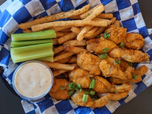 A plate of fried chicken tenders, French fries, celery sticks, and a side of ranch dressing on a blue checkered paper.