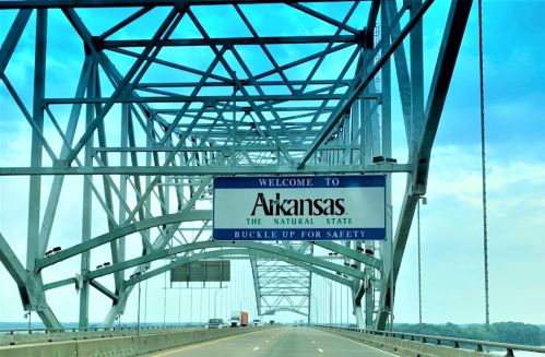 Sign welcoming drivers to Arkansas, stating "The Natural State" and "Buckle Up for Safety," with a blue sky above.