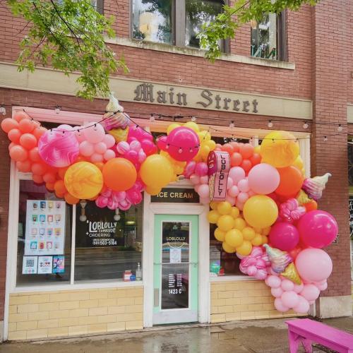 Colorful balloon arch in pink, orange, and yellow decorates the entrance of an ice cream shop on Main Street.