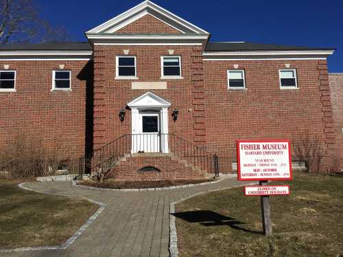 Brick building with a staircase and sign for Fisher Museum at Harvard University, surrounded by grass and blue sky.